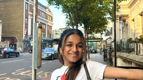 A young woman in an “I ♥ London” shirt smiles while standing at a bus stop in London, with urban streets and buildings in the background.