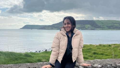 A young woman in a beige jacket sits on a stone wall with a serene coastal landscape and cloudy sky in the background in Dublin.