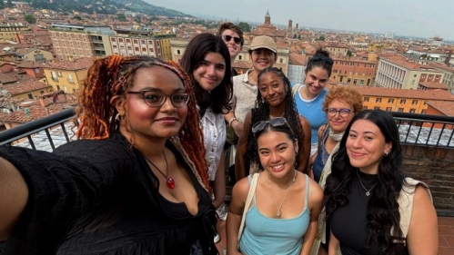 A cheerful group selfie of friends on a rooftop in Italy, with a panoramic view of the city and distant hills.