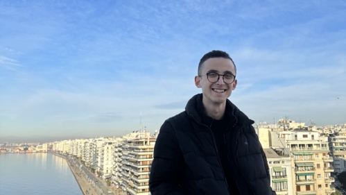 A young man in a black jacket smiles while posing against a stunning cityscape and waterfront in Thessaloniki under a bright blue sky.