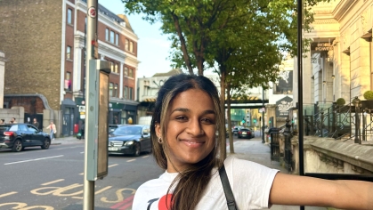 A young woman in an “I ♥ London” shirt smiles while standing at a bus stop in London, with urban streets and buildings in the background.