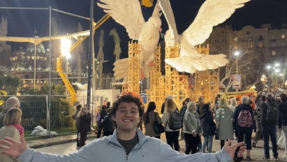 A man in a light gray sweater stands with arms wide open in front of illuminated giant dove sculptures during a nighttime festival in Valencia.