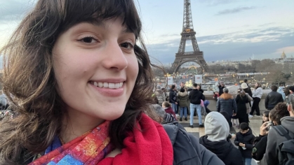 A smiling woman takes a selfie with the Eiffel Tower and a crowd of tourists in the background during a Paris evening.