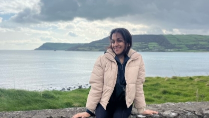 A young woman in a beige jacket sits on a stone wall with a serene coastal landscape and cloudy sky in the background in Dublin.