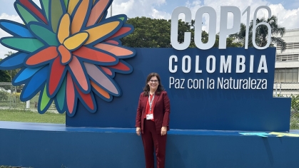 The image captures a professional moment with Pam McElwee standing confidently in front of a colorful COP16 Colombia display featuring the theme “Paz con la Naturaleza” (Peace with Nature). The vibrant backdrop emphasizes biodiversity and environmental harmony, aligning with the conference’s mission. Her attire and setting reflect an active engagement in global environmental discussions.