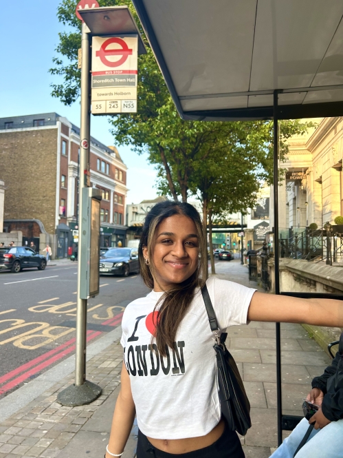 A young woman in an “I ♥ London” shirt smiles while standing at a bus stop in London, with urban streets and buildings in the background.