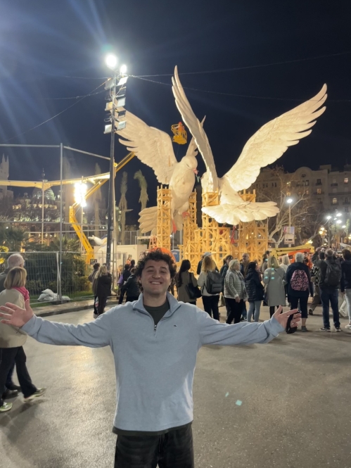 A man in a light gray sweater stands with arms wide open in front of illuminated giant dove sculptures during a nighttime festival in Valencia.