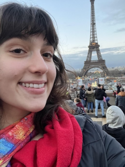 A smiling woman takes a selfie with the Eiffel Tower and a crowd of tourists in the background during a Paris evening.
