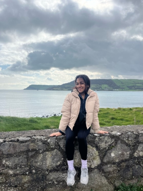 A young woman in a beige jacket sits on a stone wall with a serene coastal landscape and cloudy sky in the background in Dublin.