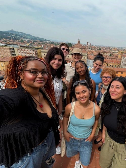 A cheerful group selfie of friends on a rooftop in Italy, with a panoramic view of the city and distant hills.