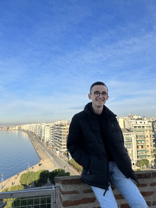 A young man in a black jacket smiles while posing against a stunning cityscape and waterfront in Thessaloniki under a bright blue sky.