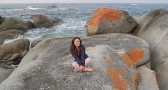 Claire sits cross-legged on a large boulder with orange algae on the coast of the ocean