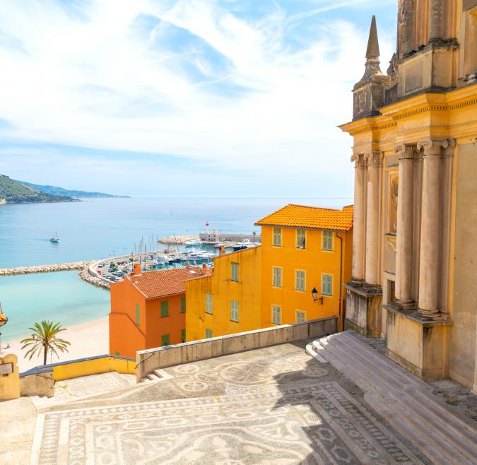 Standing over a mosaic staircase, the ocean and some mountains are visible and it is sunny. There are balconies with potted plants and terra cotta roofs. 