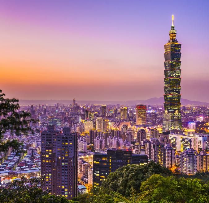 From a hilltop, the Taipei skyline is viewed. There are many skyscrapers, but one is towering over the rest. It is sunset, so all is illuminated orange and purple