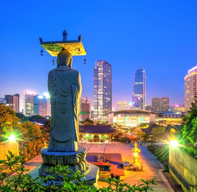 A statue of Buddha is illuminated at night, overlooking the Seoul skyline